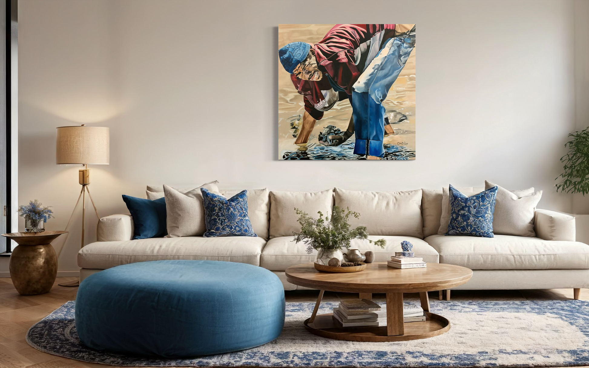 Living room with a beige sofa, blue ottoman, and decorative pillows and artwork on the wall of a man with his hand in water, looking for oysters to sell.